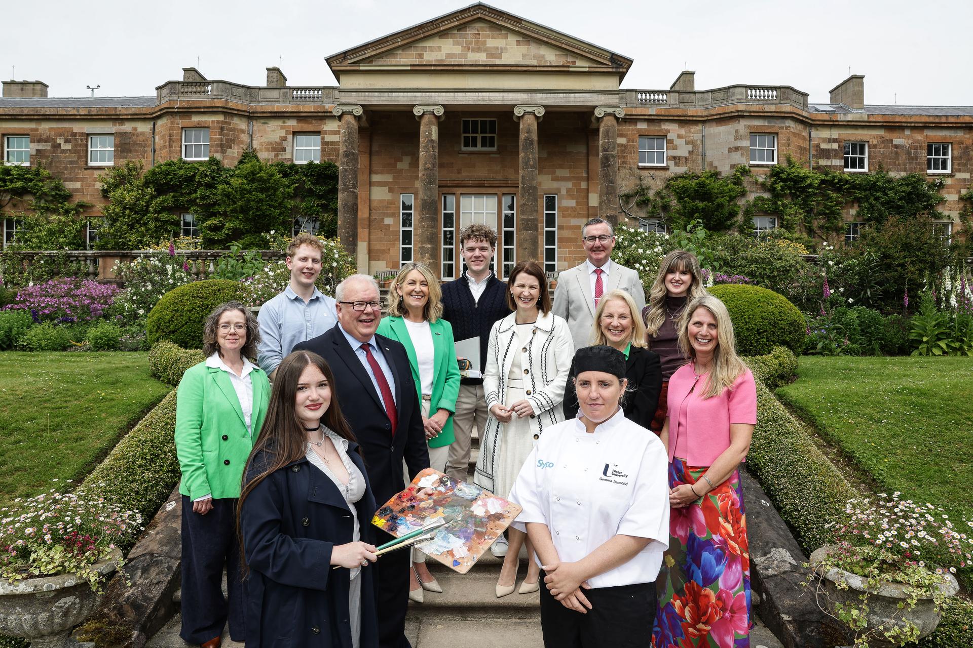 Group photograph of thirteen people standing on the steps of a formal garden in front of a large historic stone building with columns. The group includes individuals in business attire alongside people holding an artist&rsquo;s palette and wearing a chef&rsquo;s uniform, surrounded by colourful flower beds and manicured lawns.